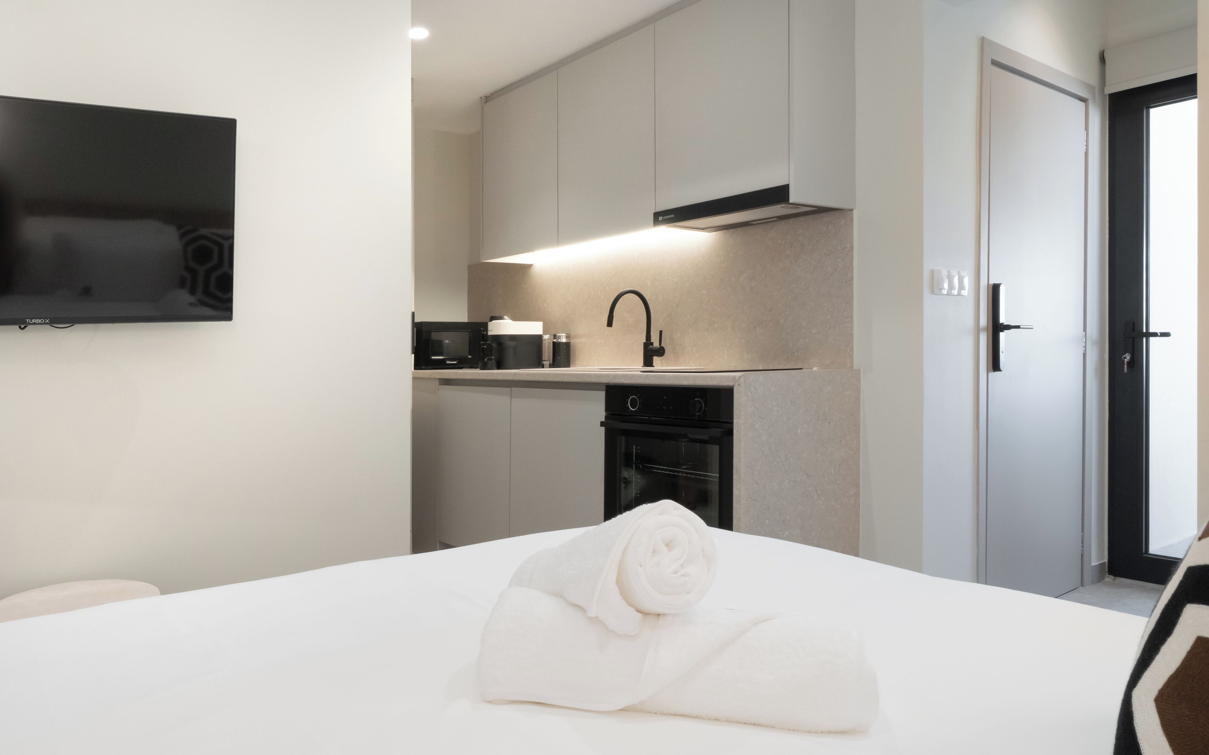Une chambre d’hôtel moderne équipée d’un lit, d’une serviette blanche soigneusement pliée, d’un four à micro-ondes encastré, d’un lavabo et d’un téléviseur fixé au mur.