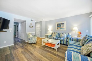 Een woonkamer met een bank, fauteuil en salontafel, een aan de muur bevestigde televisie en een deur naar een andere kamer.