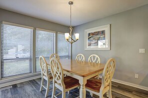 A dining room with a wooden table, six chairs, a chandelier, and large windows with blinds.