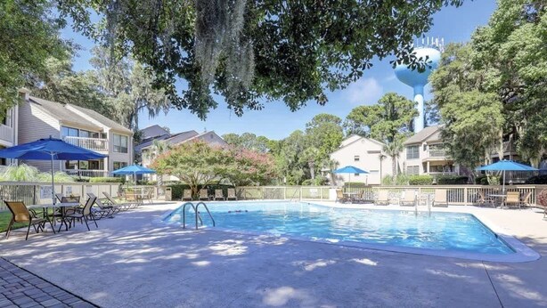 Una piscina con sillas de descanso y sombrillas, rodeada de casas y árboles.