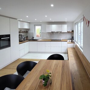 A modern kitchen with white cabinets, a wooden dining table, and a window providing natural light.