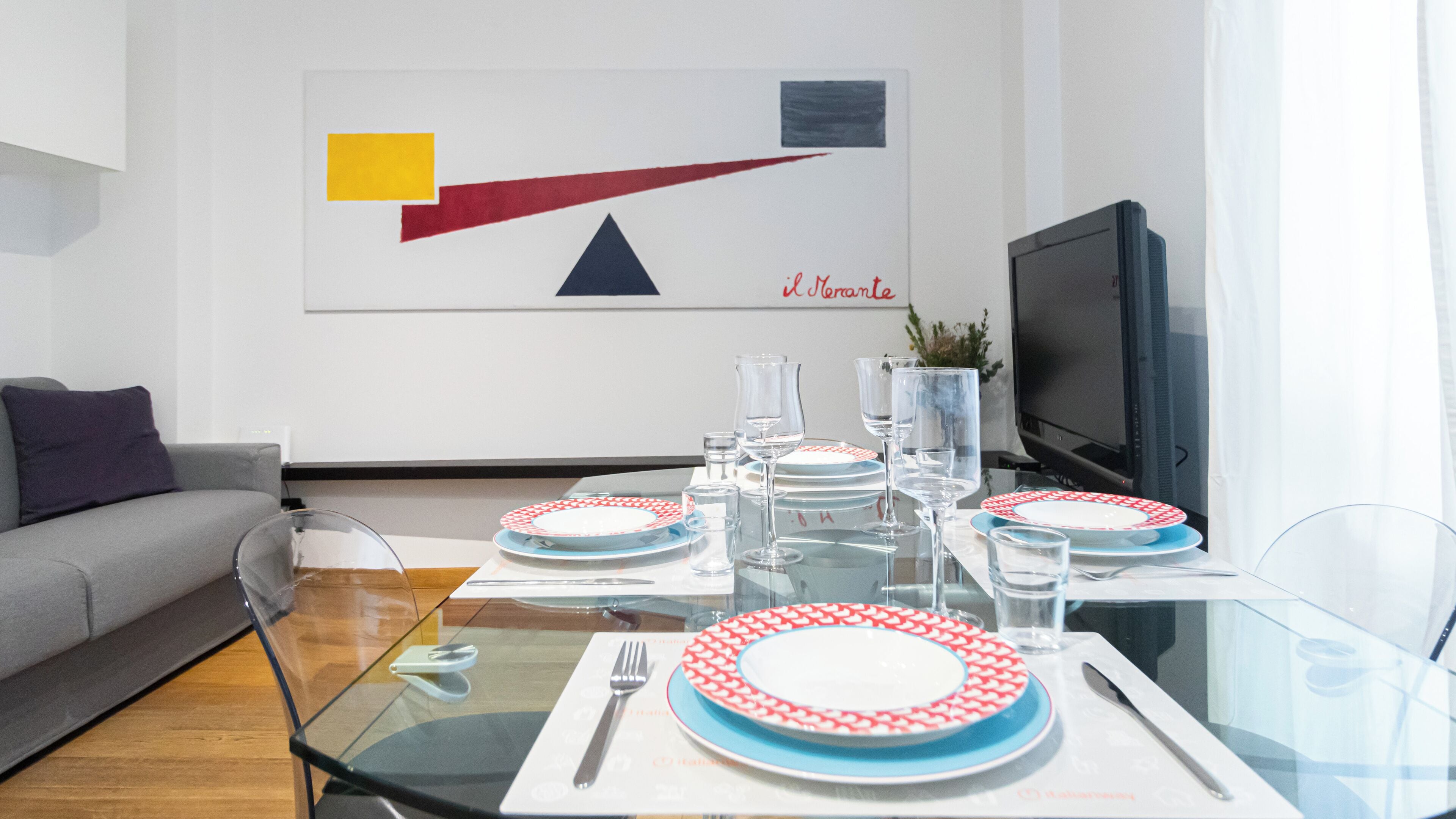 A modern living room with a glass dining table set for four, a sofa, a flat-screen TV, and a wall-mounted abstract painting.