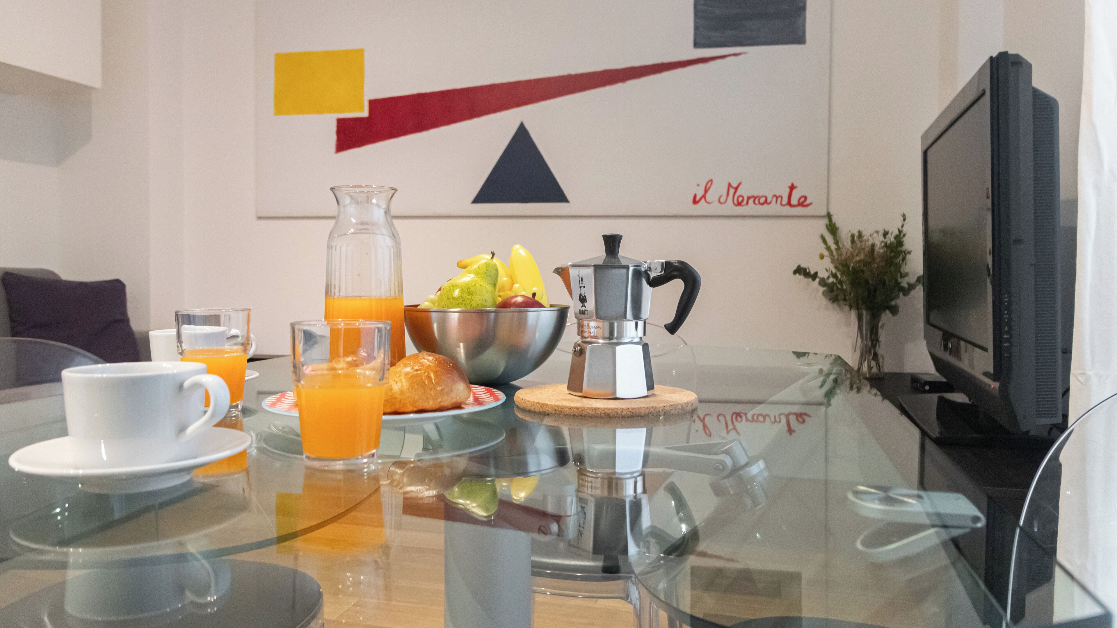 Una mesa de desayuno con cafetera, un tazón de fruta y vasos de jugo de naranja.
