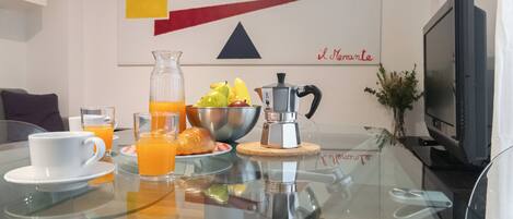 Una mesa de desayuno con cafetera, un tazón de fruta y vasos de jugo de naranja.