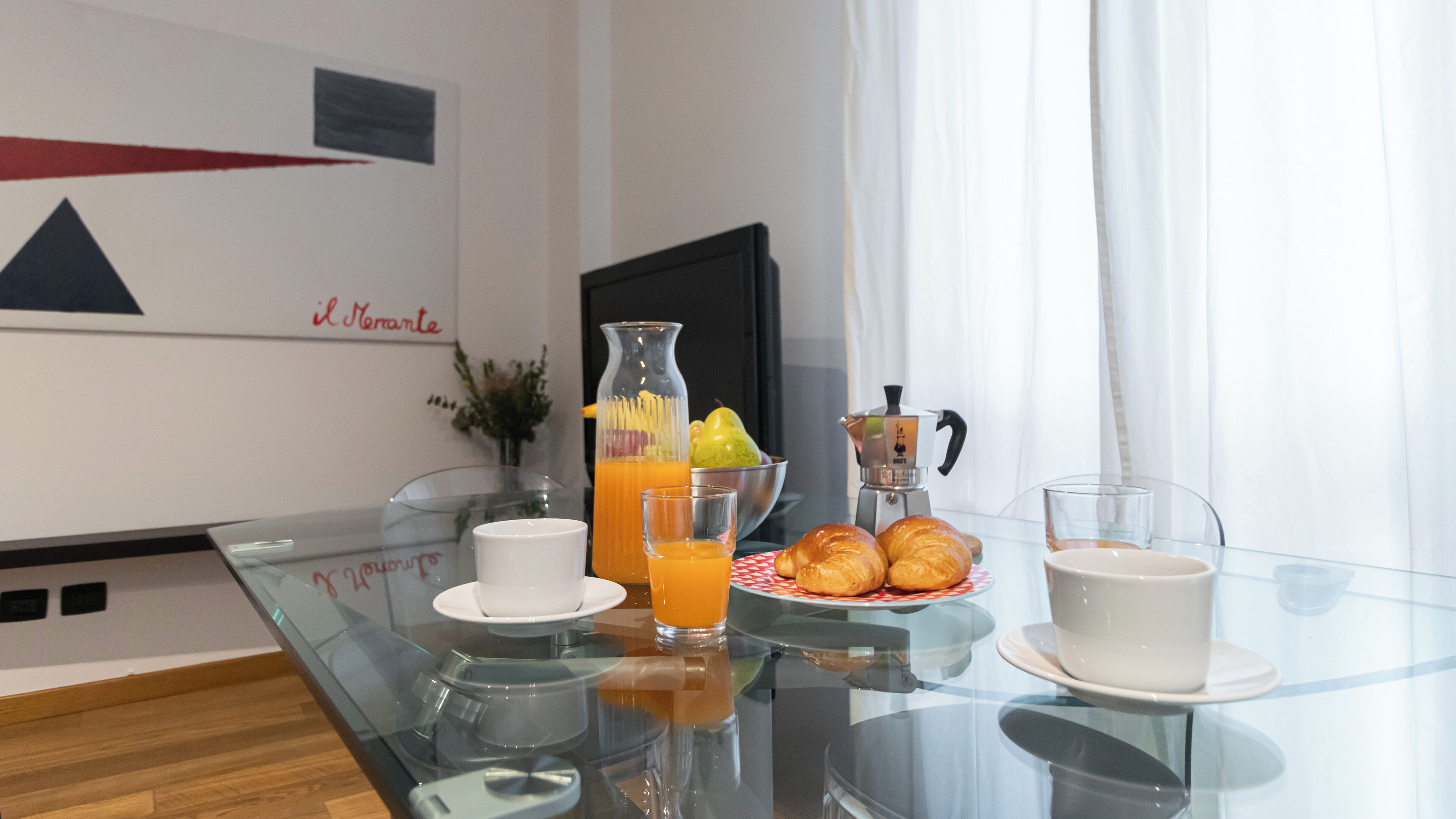 Een ontbijt met croissants, een schaal fruit, een koffieapparaat en een glas jus d'orange op een glazen tafel.