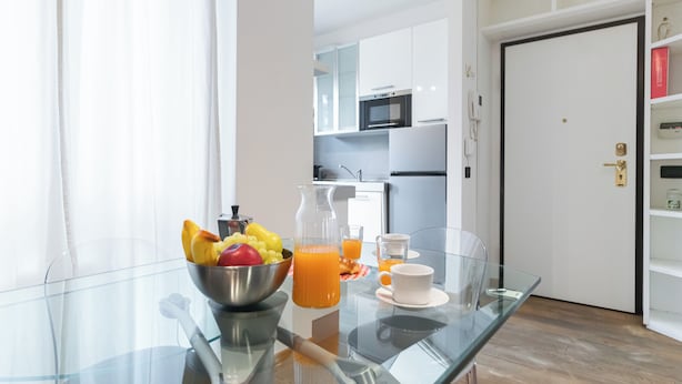 A modern kitchen with white cabinets, a microwave, and a refrigerator. A glass table holds a fruit bowl, a pitcher of orange juice, and a cup. - Italianway Comfort - Gioia 59 (Milan)