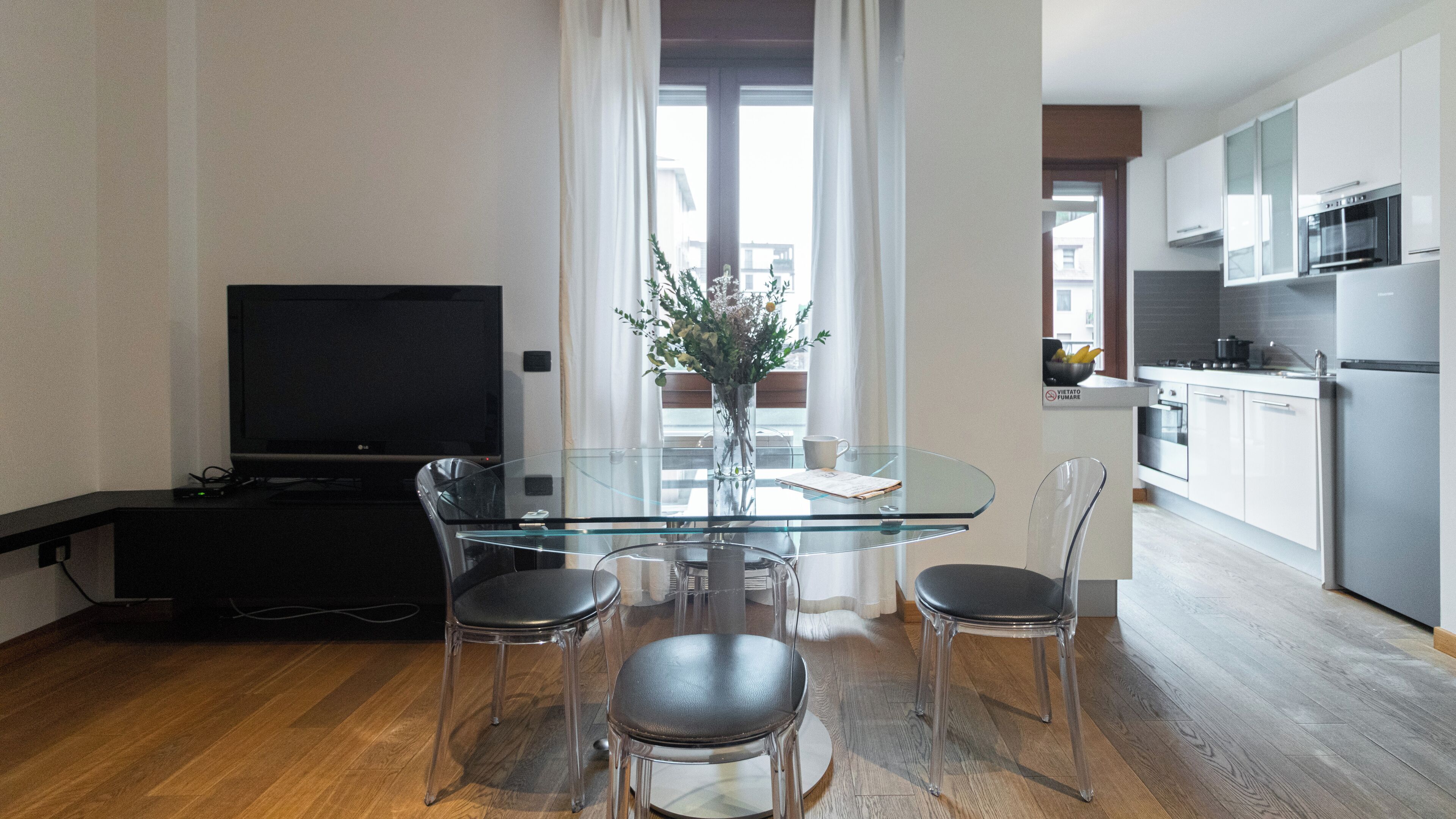 Un espacio habitable moderno con una mesa de vidrio, dos sillas, un televisor y una zona de cocina.
