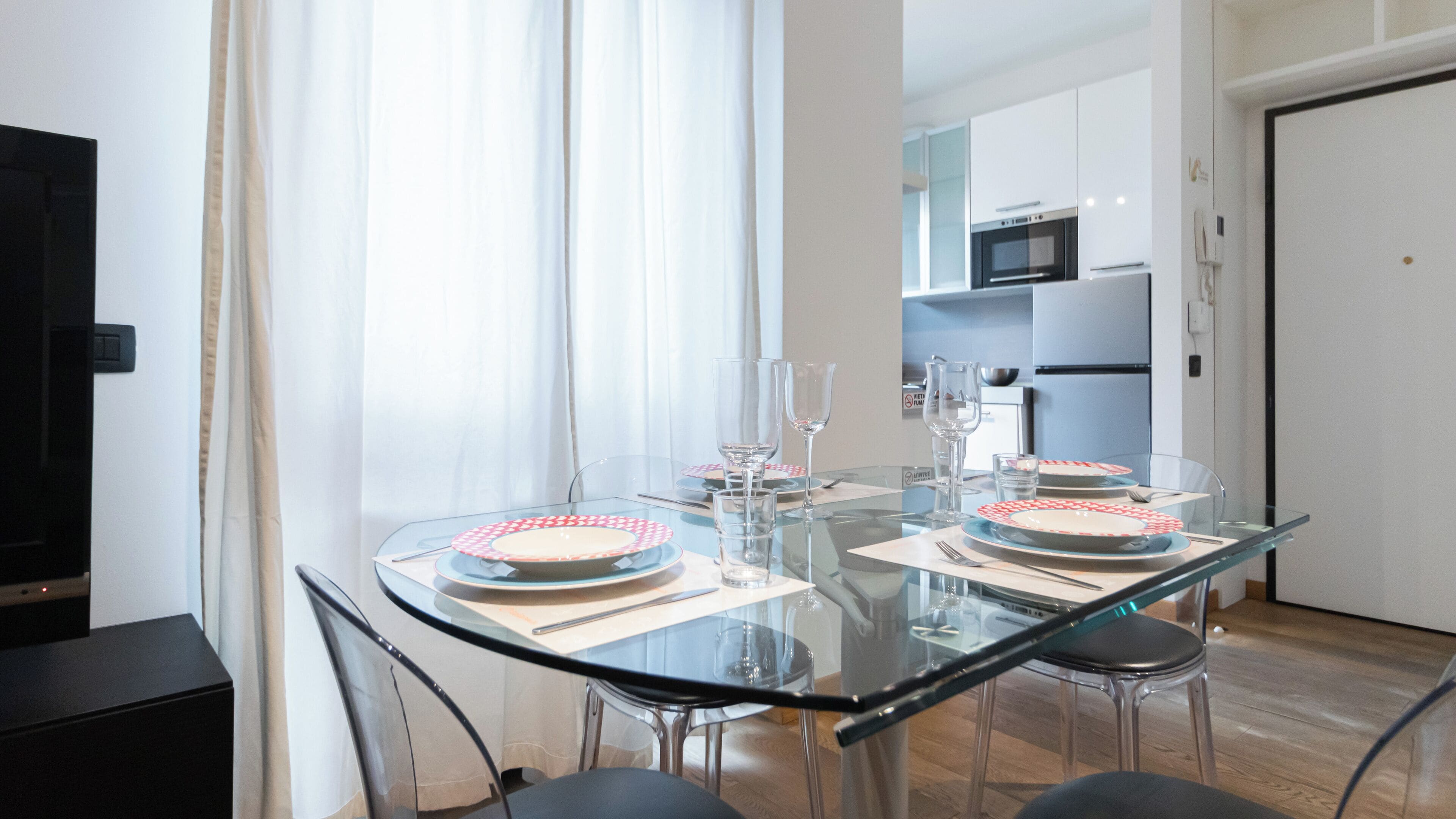 A modern dining area with a glass table set for two, surrounded by chairs. A kitchen with white cabinets and appliances is visible in the background.