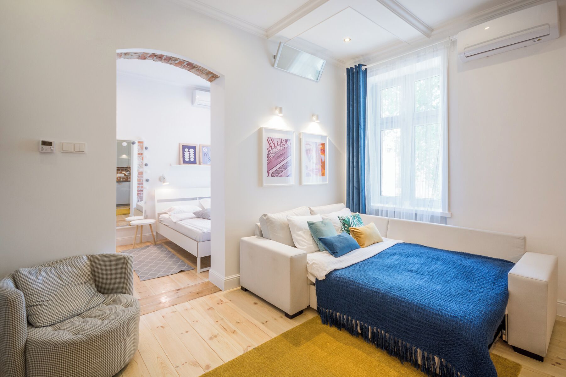 Ein modernes Schlafzimmer mit Bett, einer Couch und einem Sessel. Ein Fenster mit Vorhängen und eine Wand-Klimaanlage sind vorhanden.