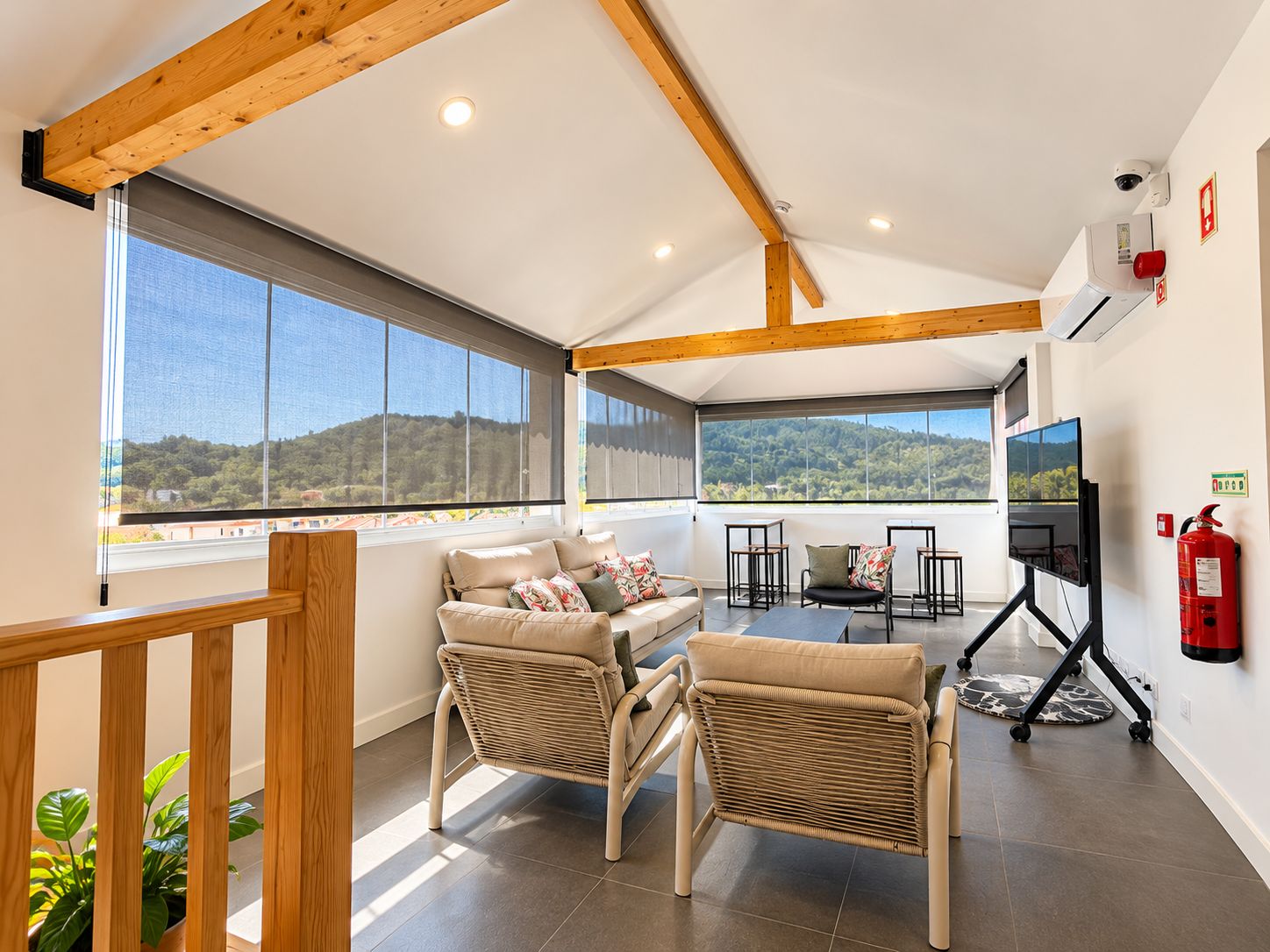 A spacious room with a high ceiling, large windows, a sofa, chairs, a TV, and a fire extinguisher.