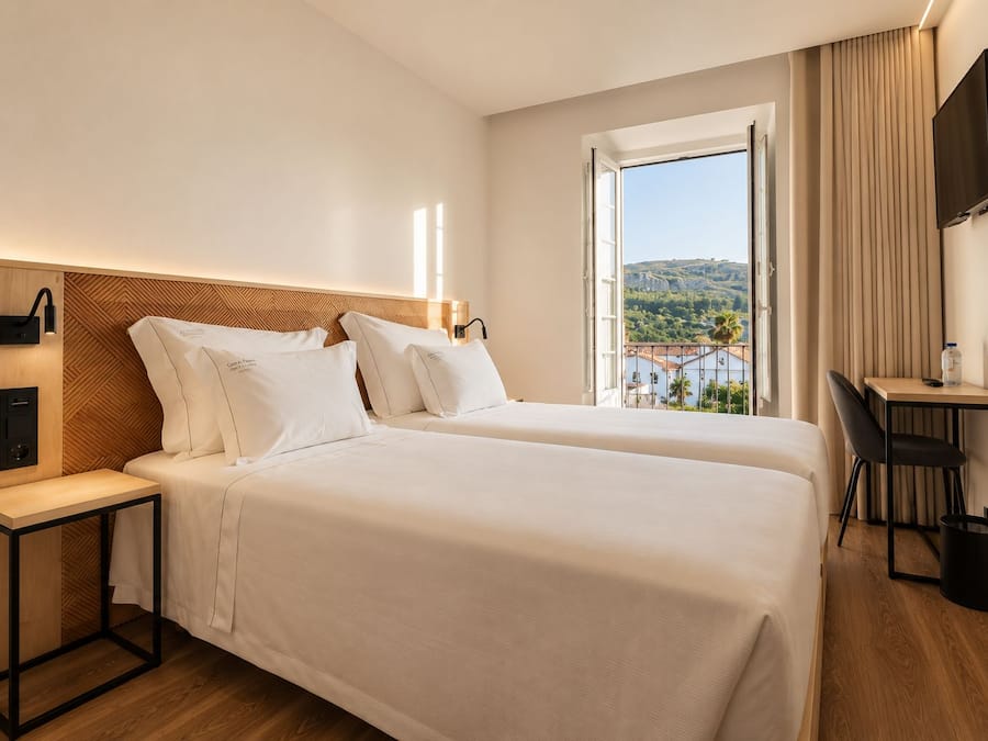 A hotel room with two beds, a desk, a chair, a TV, and a window with a view.