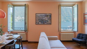 A room with a dining table set for four, a sofa, a chair, and a large window with a view of a body of water and hills. - Cernobbio in Riva. Lake views, Steps from Lake. (Cernobbio)