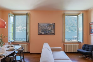 A room with a dining table set for four, a sofa, a chair, and a large window with a view of a body of water and hills. - Cernobbio in Riva. Lake views, Steps from Lake. (Cernobbio)