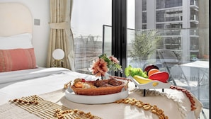 Quarto de hotel com uma cama, bandeja de café da manhã com frutas e doces, um vaso de planta e vista para a cidade.