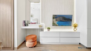 Uma sala de estar moderna com uma mesa consola branca, uma TV montada exibindo uma paisagem costeira e um vaso com planta.
