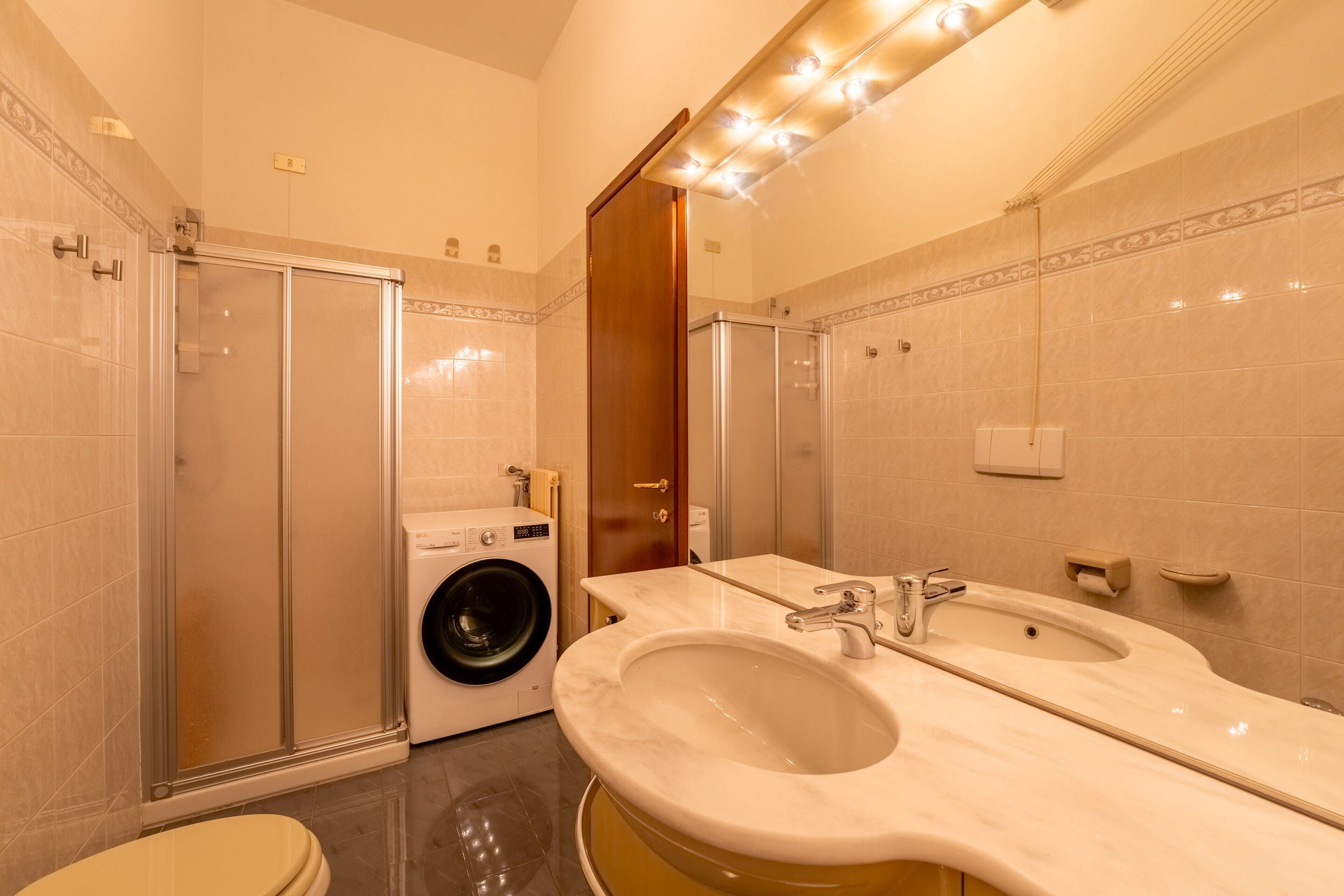A modern bathroom with a glass shower enclosure, a built-in washing machine, and a double sink vanity.