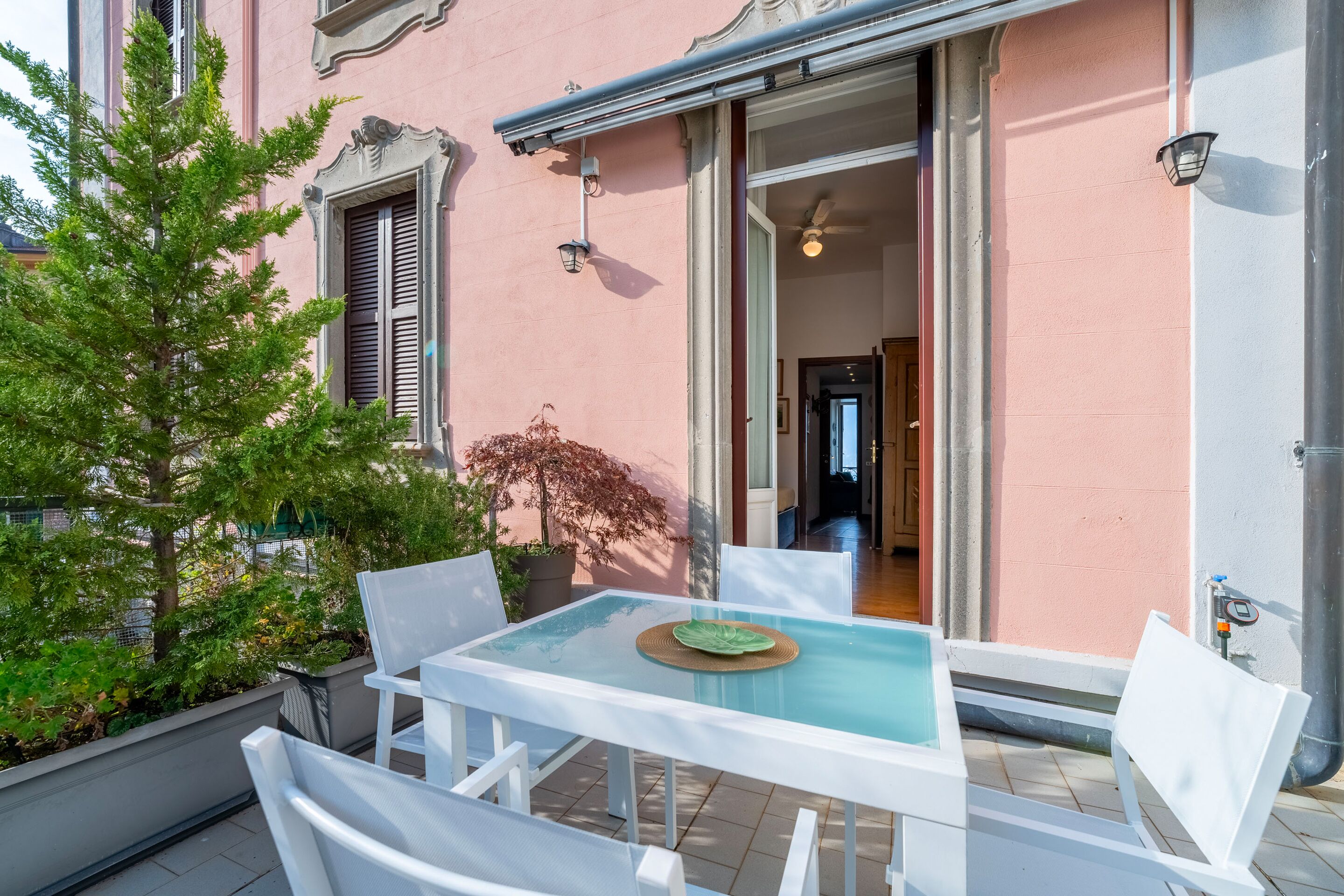 Une table à plateau en verre, des chaises blanches, une plante en pot et une vue sur un bâtiment rose avec une porte et une fenêtre.
