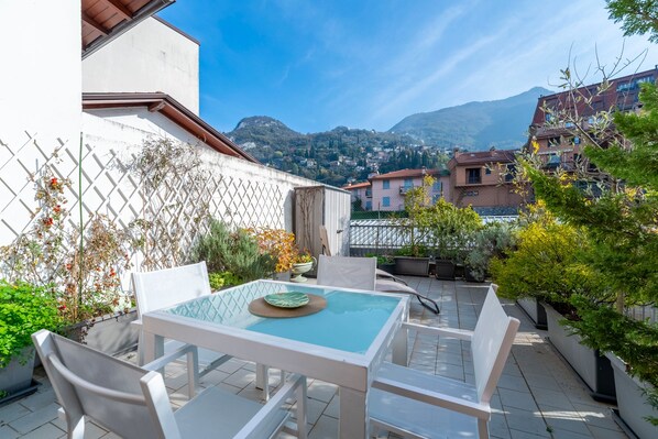 Patio esterno con tavolo e sedie, circondato da piante in vaso e vista su una montagna.