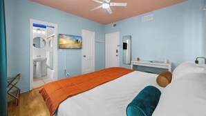 A bedroom with a bed, a ceiling fan, a TV, a mirror, a nightstand, and a bathroom with a sink and mirror. - #05 Corner Unit in Mid-century Art-Déco Building KingBed EnSuiteBath Steps>Beach (Miami Beach)