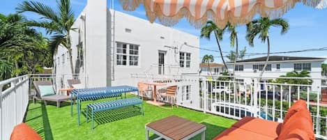 A white building with a fenced patio area, outdoor seating, and palm trees.