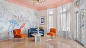 Une pièce avec un plancher ciré, un canapé bleu, deux fauteuils orange, une table basse blanche ornée de fleurs, une fresque murale représentant des oiseaux et un lustre.