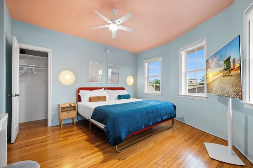 Quarto com cama, mesa de cabeceira, ventilador de teto e janela vista para a praia.
