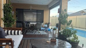 Une terrasse extérieure couverte, avec une table ronde, des coussins à rayures et une piscine en arrière-plan.