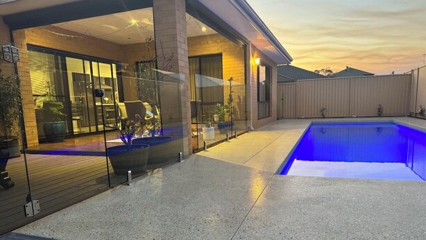 Un espace piscine extérieur moderne, doté d’une structure en verre, d’une terrasse en bois et d’un patio en béton.