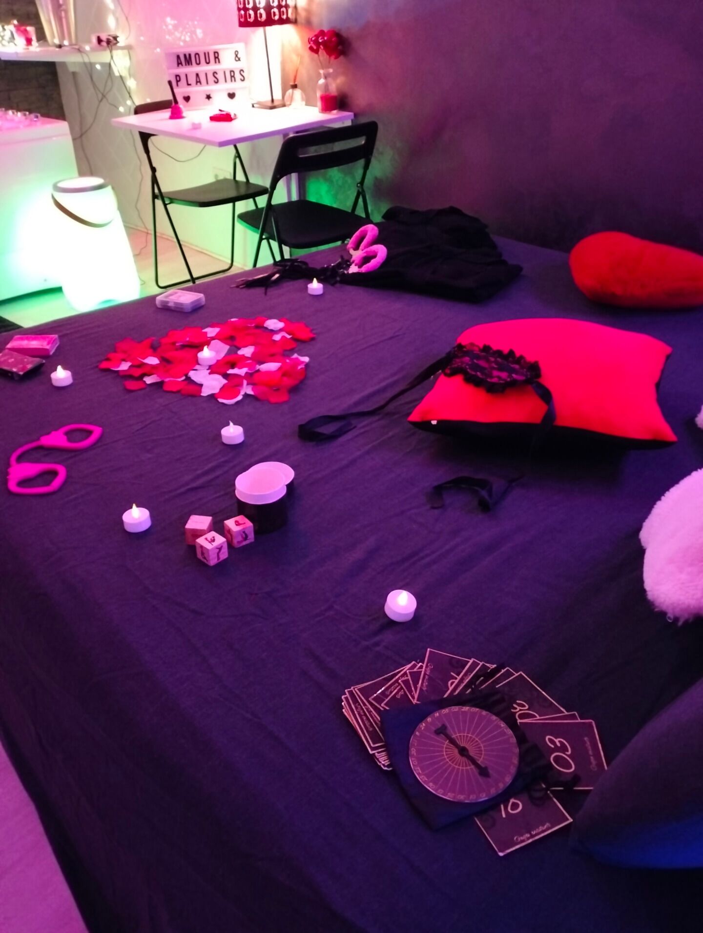 A room with a bed, a table, and a chair. There are candles, a clock, and a sign that says "AMOUR & PLAISIR".