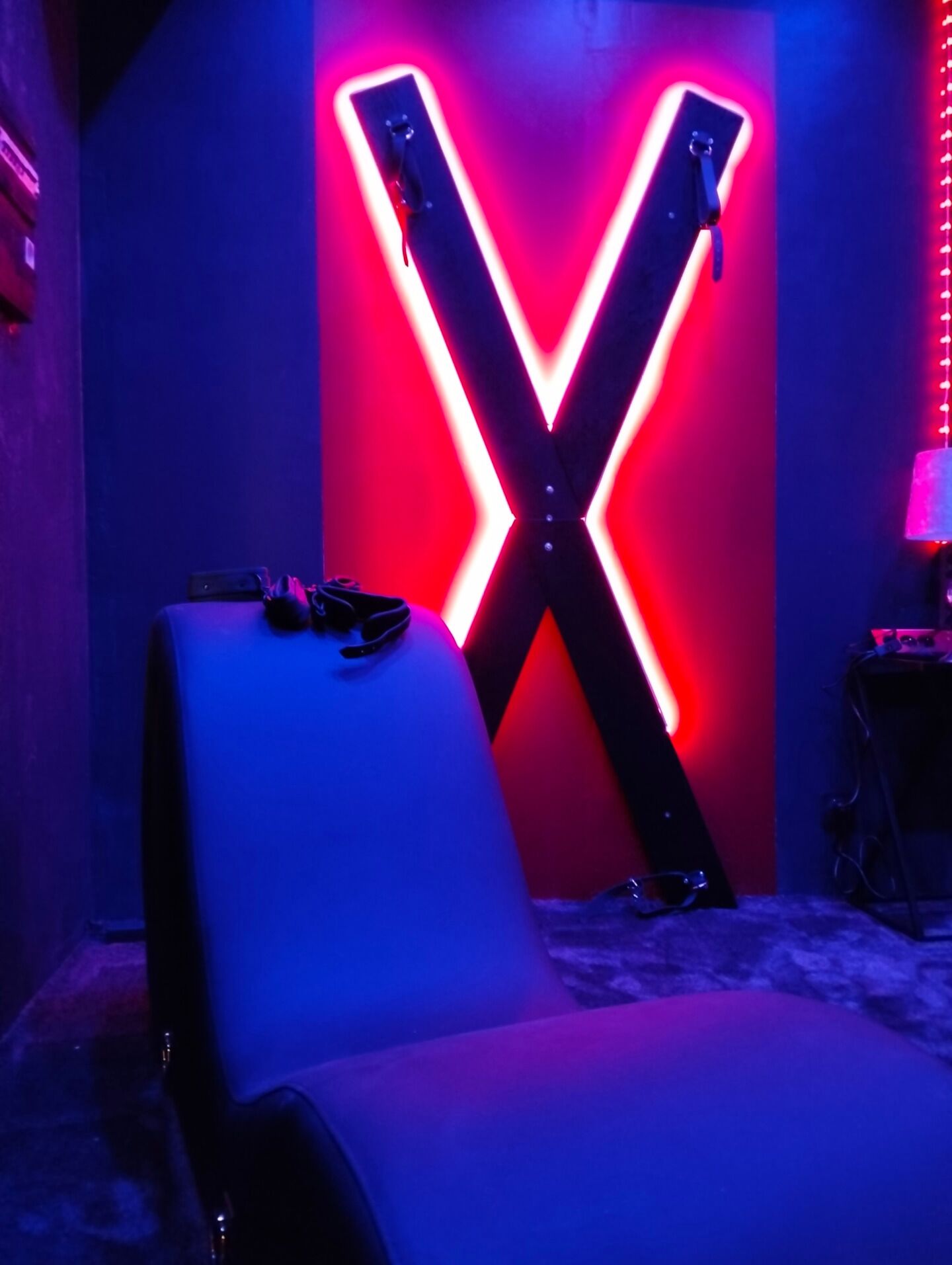 Een kamer met een grote, verlichte X op de muur, een witte stoel en een kleine tafel met spullen erop.