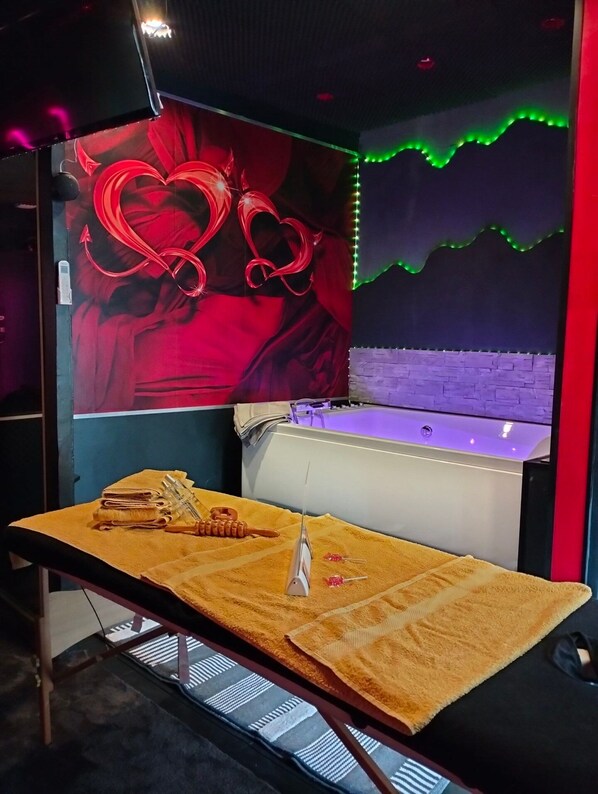 A massage table with towels and a massage gun, a wall with a heart design, and a brick wall with green LED lights. - Loveroom Toulon (Toulon)