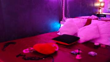 Una habitación con una cama, una manta roja, un almohadón blanco y un bolso rojo. También hay varios objetos sobre la cama, incluyendo una billetera negra y un objeto rosa.