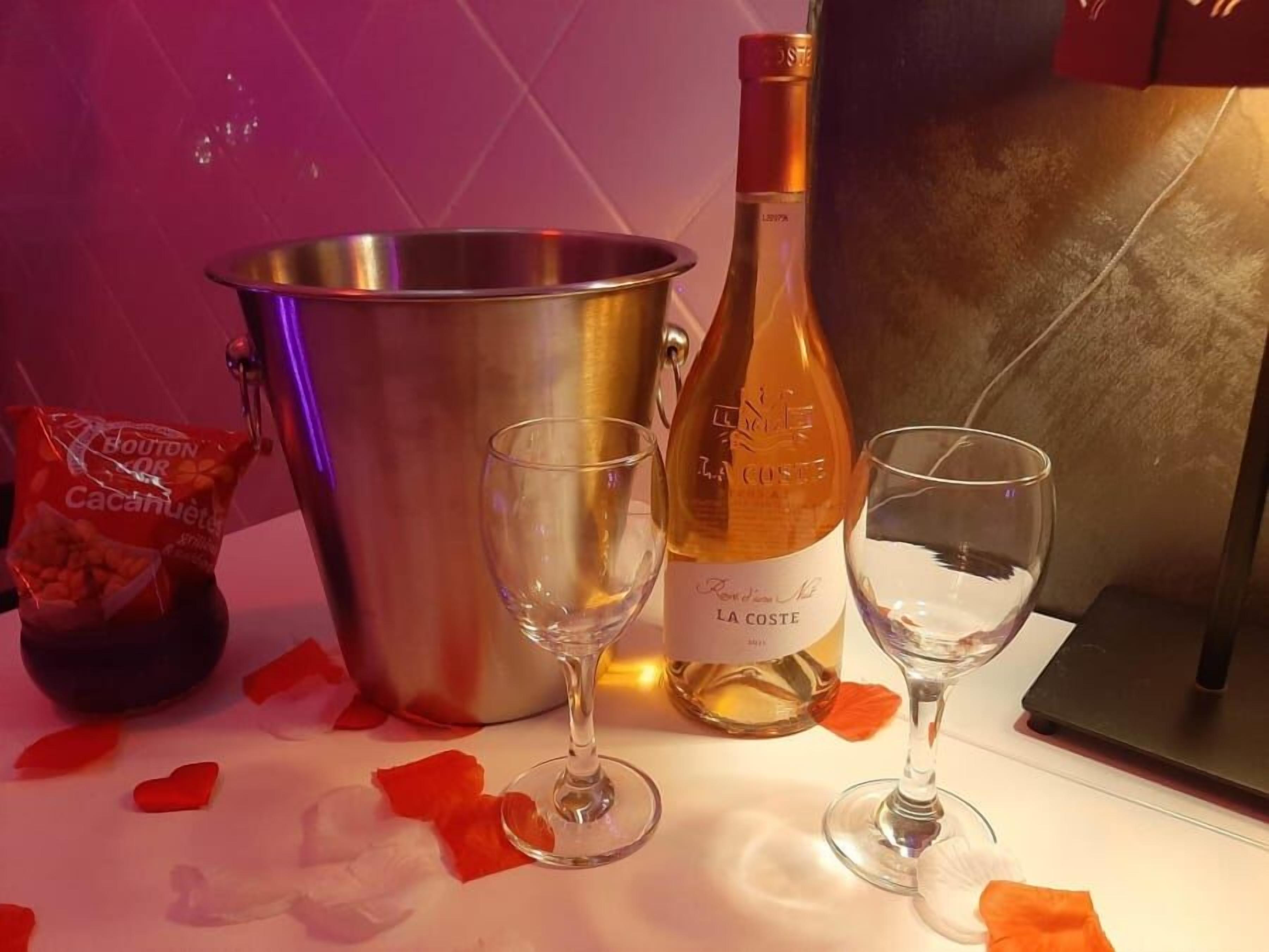 Una botella de vino, dos copas de vino, un balde de metal y una bolsa de aperitivos sobre una mesa con pétalos de rosa esparcidos.