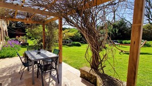 A covered patio with a table and chairs, surrounded by greenery and a garden. - Les Granges Indigo (Fossemagne)