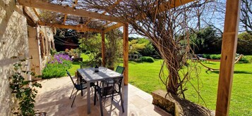 A covered patio with a table and chairs, surrounded by greenery and a garden.