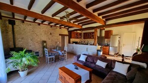 A spacious living area with a stone wall, wooden beams, a dining set, a sofa, and a kitchen in the background. - Les Granges Indigo (Fossemagne)