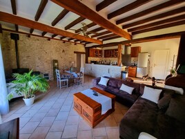 A spacious living area with a stone wall, wooden beams, a dining set, a sofa, and a kitchen in the background.