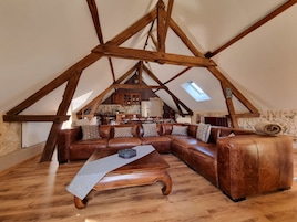 A loft-style living space with a wooden ceiling structure, a brown leather sectional sofa, a wooden coffee table, and a stone wall.