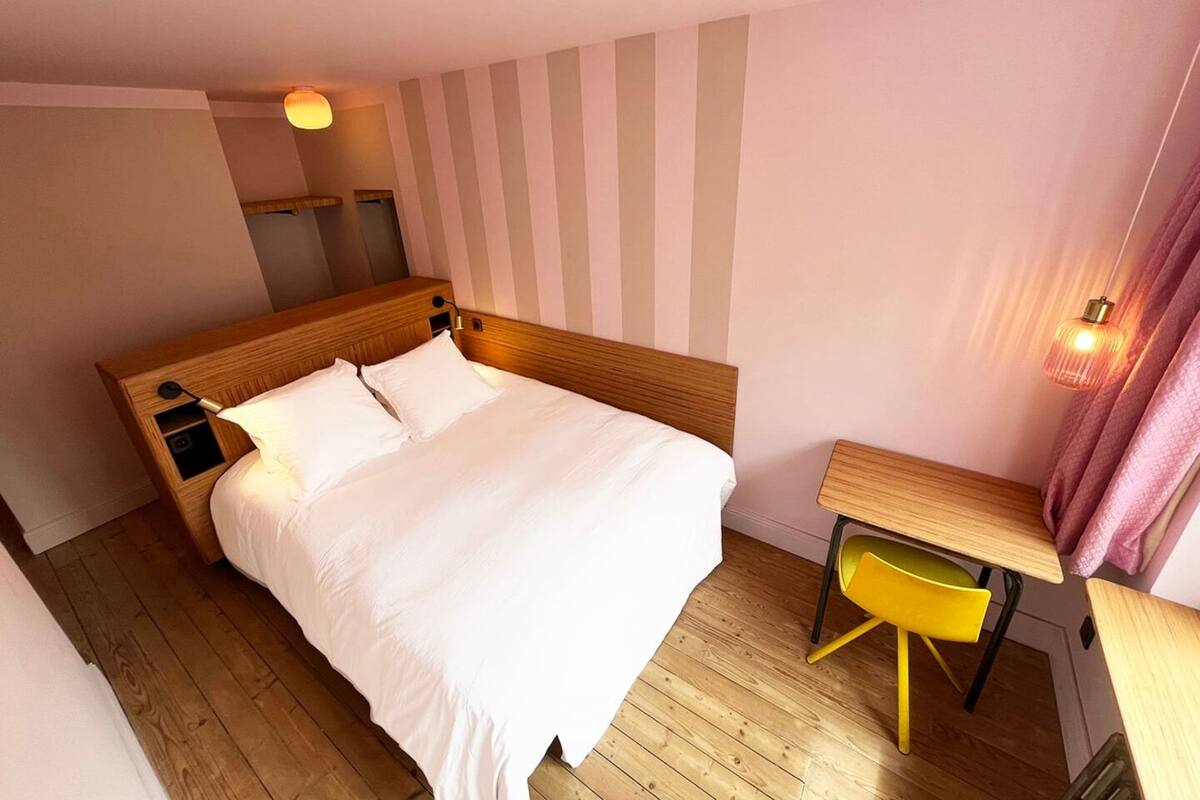 A hotel room with a wooden bed, a small desk, a chair, and a wall-mounted lamp.