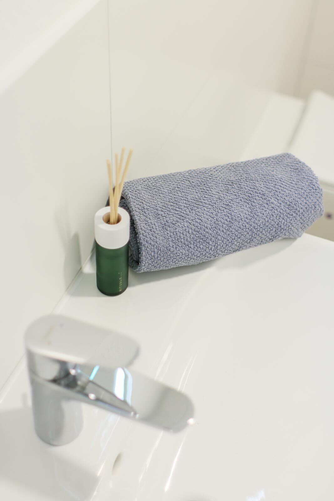 Une salle de bain avec un lavabo blanc, un récipient vert contenant des diffuseurs à roseaux et une serviette enroulée posée sur le bord.