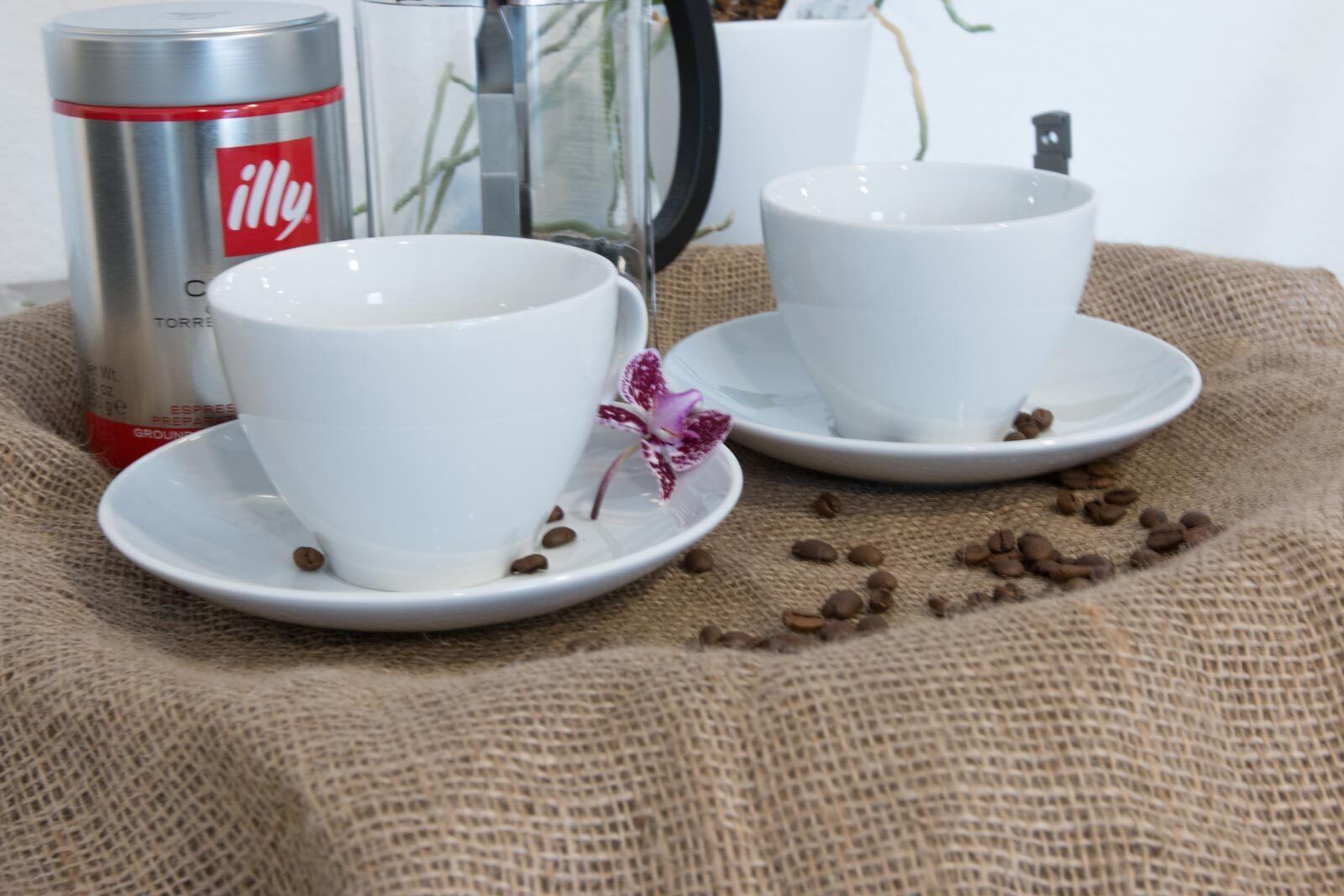 Två vita koppar på små fat, med kaffebönor och en blomma på en yta av säckväv.