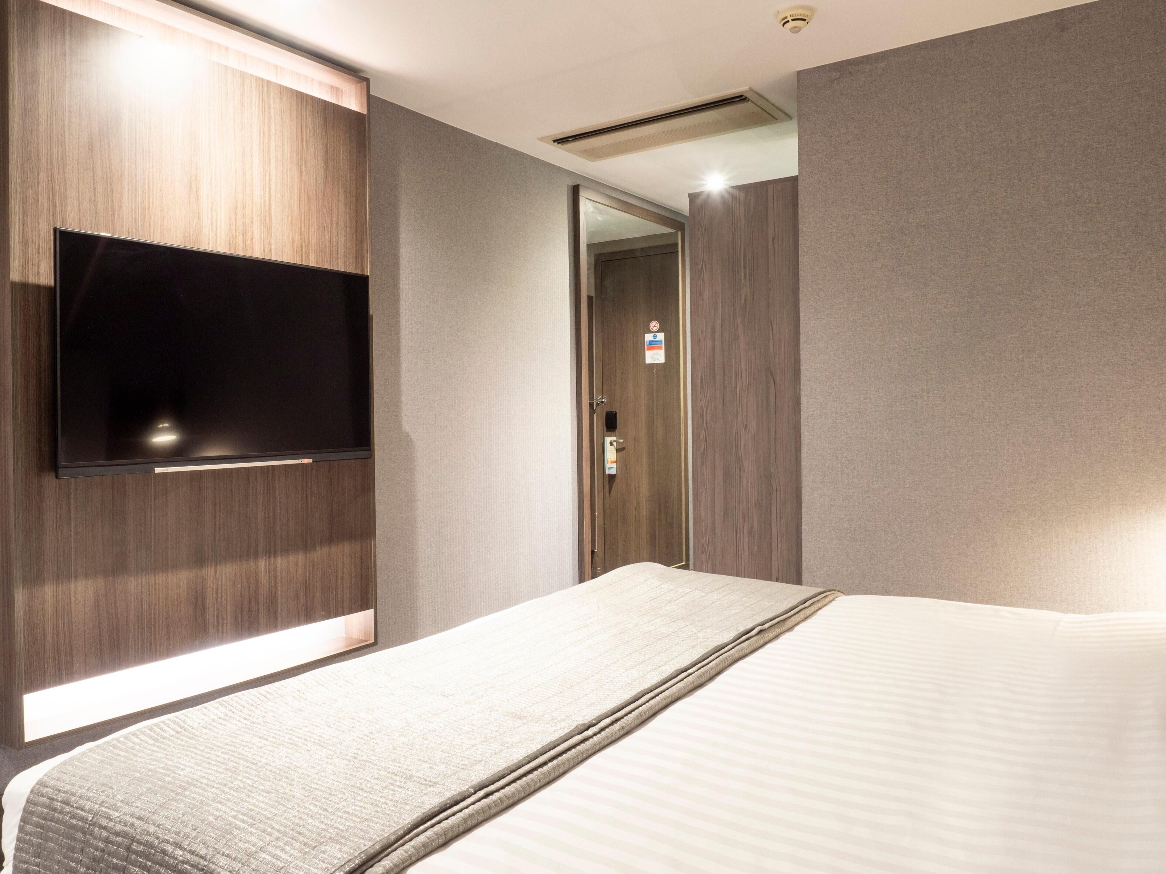 A hotel room with a bed, a flat-screen TV mounted on the wall, a door with a sign, and an air vent.