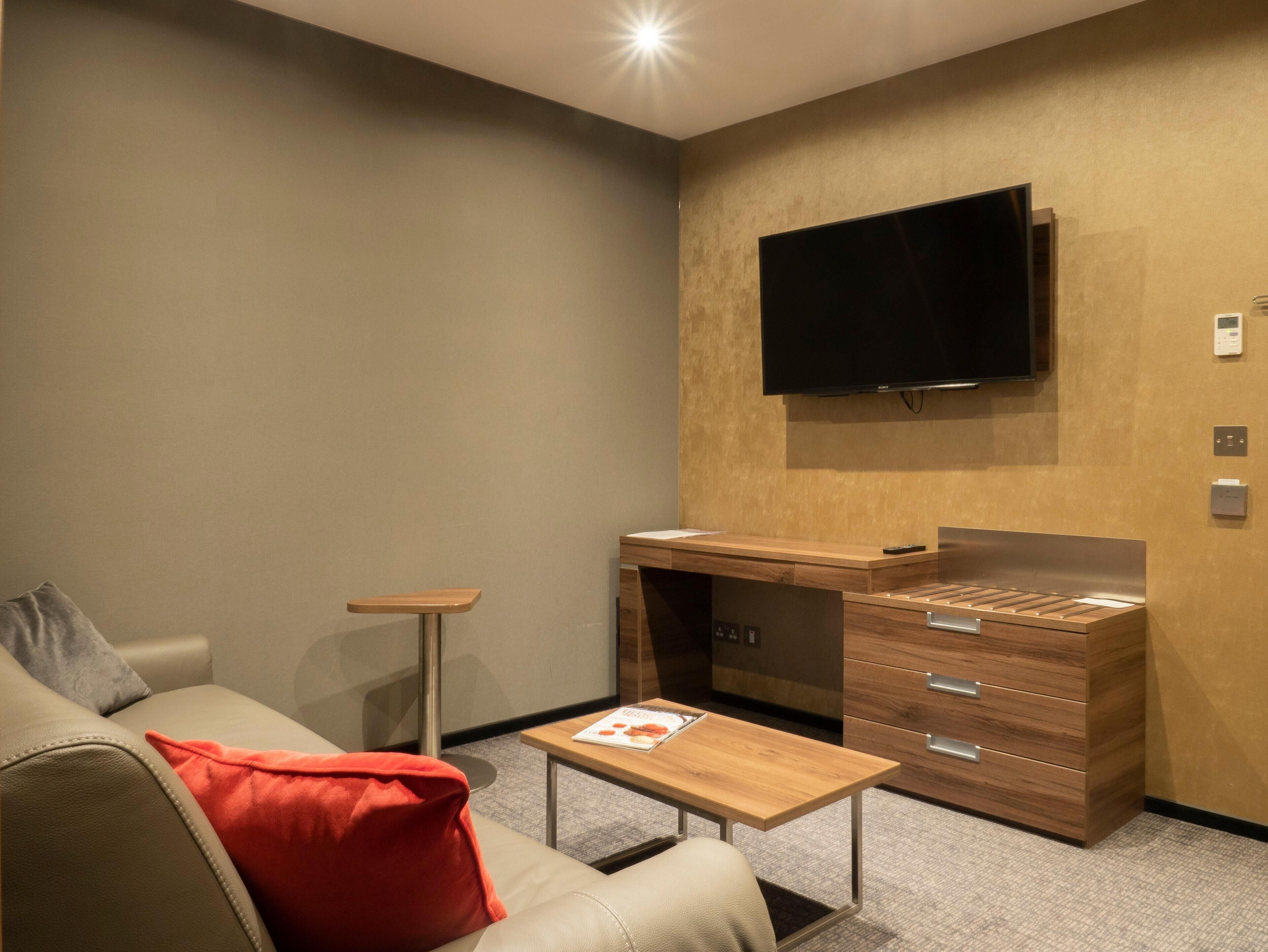 A modern hotel room with a sofa, a small wooden table, a desk with drawers, a flat-screen TV mounted on the wall, and a carpeted floor.