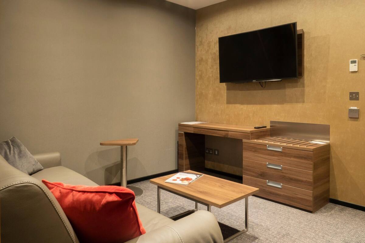 Una habitació d'hotel moderna amb un sofà, una taula de fusta petita, un escriptori amb calaixos, un televisor de pantalla plana fixat a la paret i un sòl amb moqueta.