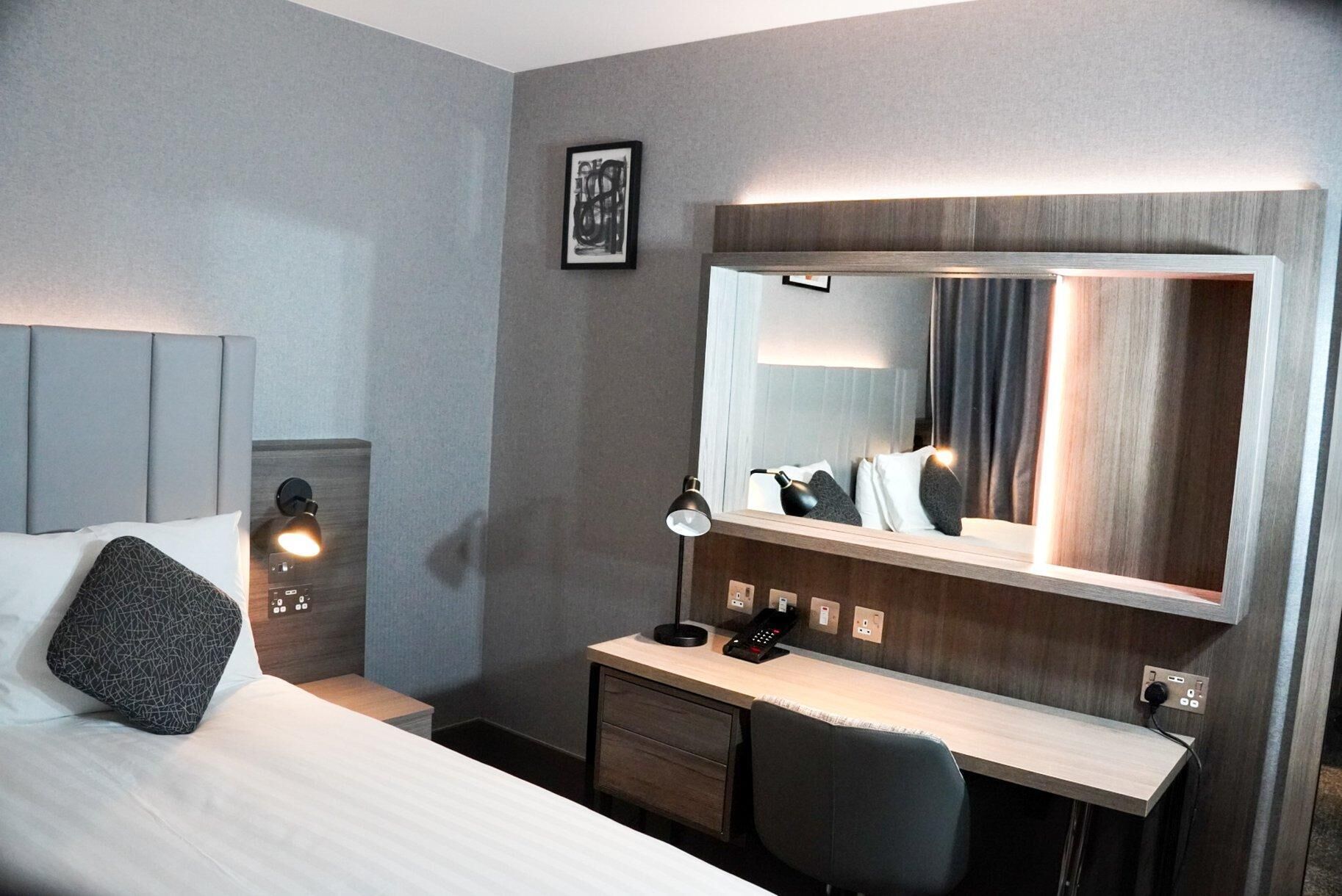 A hotel room with a bed, bedside table, desk, chair, and a large mirror.