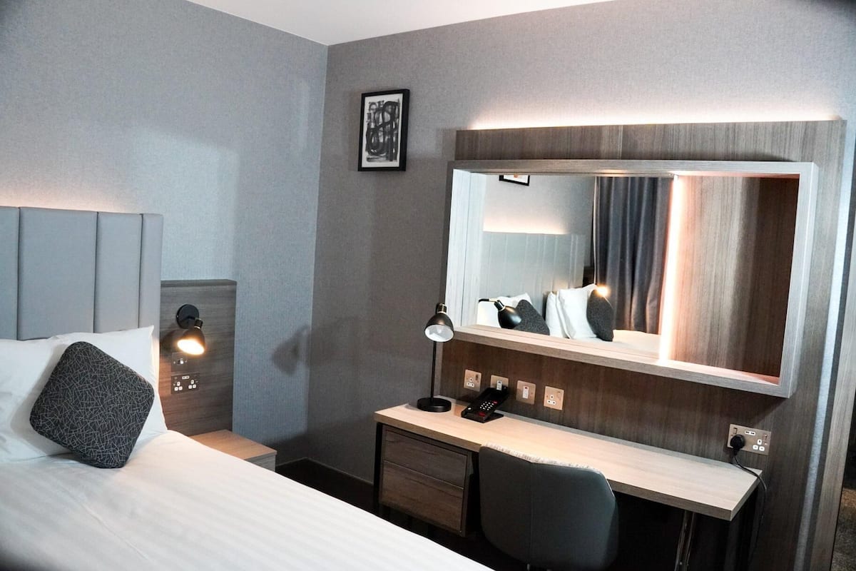 Habitació d'hotel amb llit, taulell de nit, escriptori, cadira i un mirall gran.