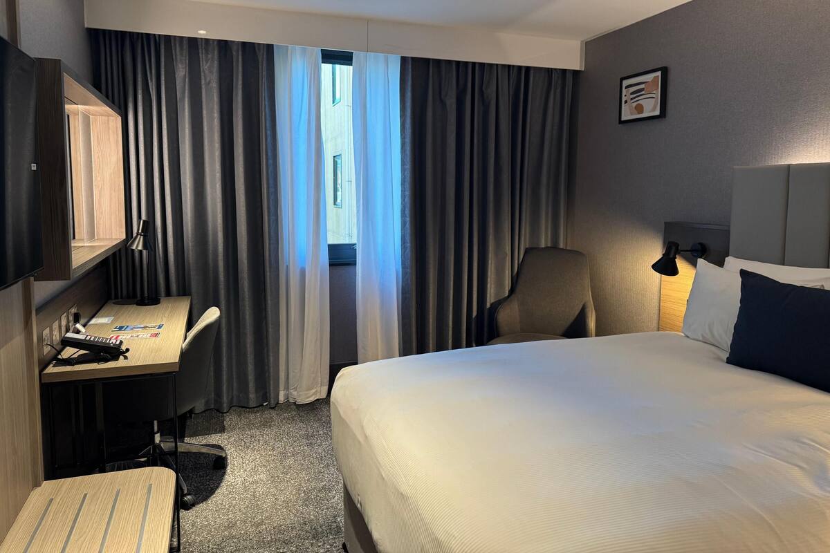 Habitació d'hotel amb llit, escriptori, cadira, televisió i finestra amb cortines.