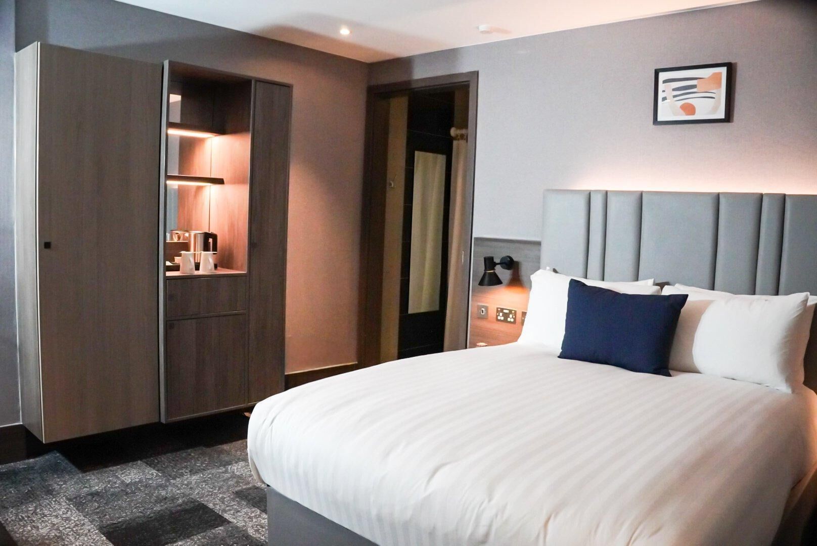 A hotel room with a large bed, a wardrobe, a bedside table with a lamp, and a wall-mounted artwork.