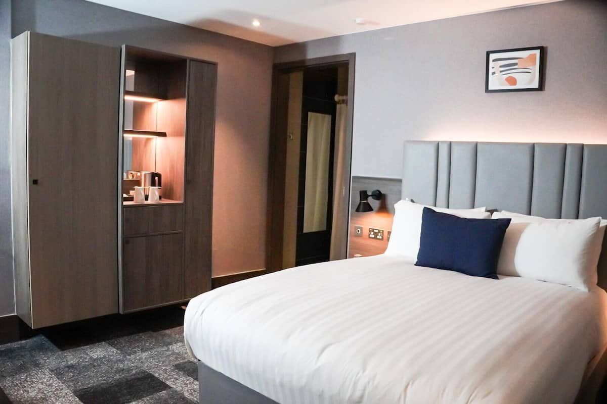 Habitació d'hotel amb un llit gran, un armari, una tauleta de nit amb una làmpada i una obra d'art penjada a la paret.