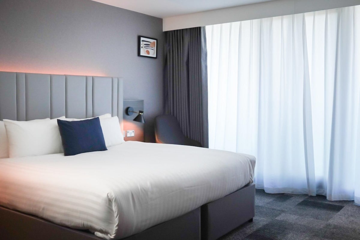 A hotel room with a large bed, a nightstand, a lamp, and a view of the outside through sheer curtains.
